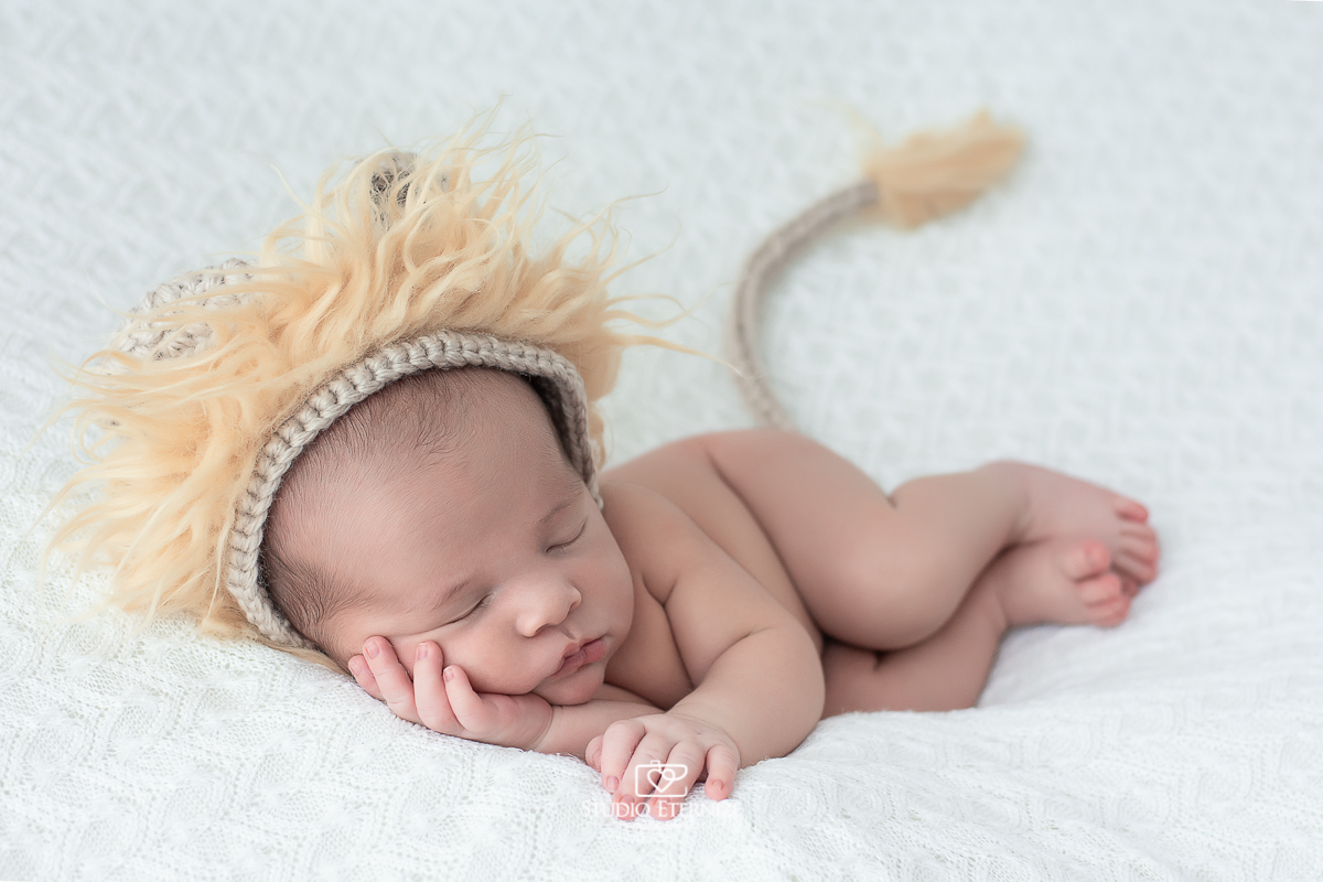 Fotografia newborn de menino realizada na cidade de Poços de Caldas - MG, pelo Studio Eternize. Bebê peladinho de touca de leão deitado de lado. 