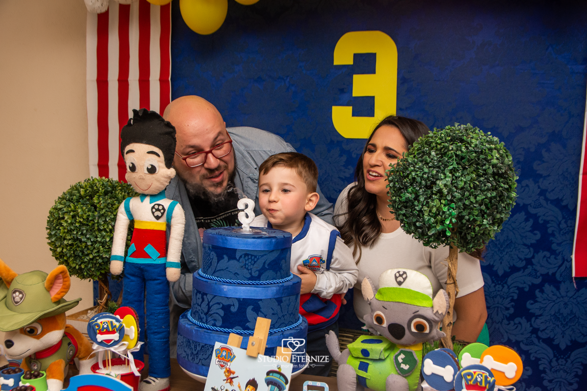 Fotografia de festa infantil de 3 anos registrada pelo Studio Eternize no tema da Patrulha Canina em no Buffet Cata-Vento em Poços de Caldas - MG. Aniversariante e seus pais soprando a vela do bolo.