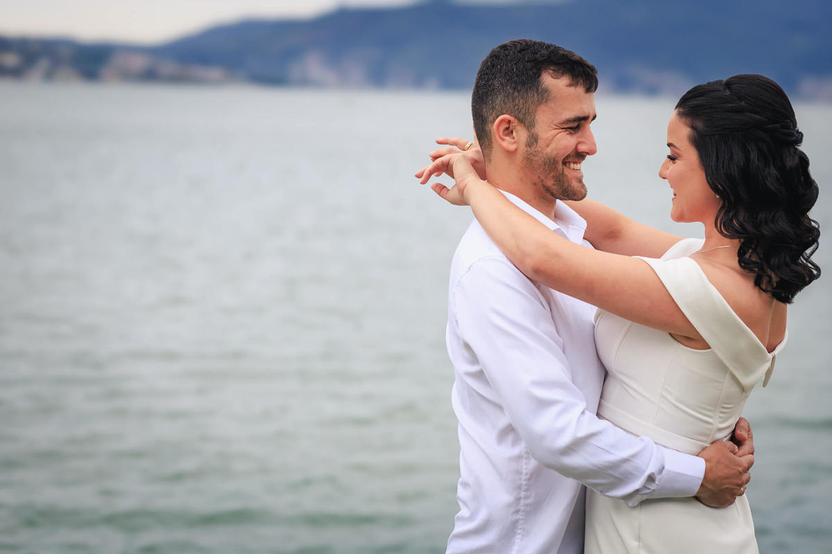 Djonata e Aline trocando olhares apaixonados em um cenário romântico à beira-mar em Coqueiros, Florianópolis.