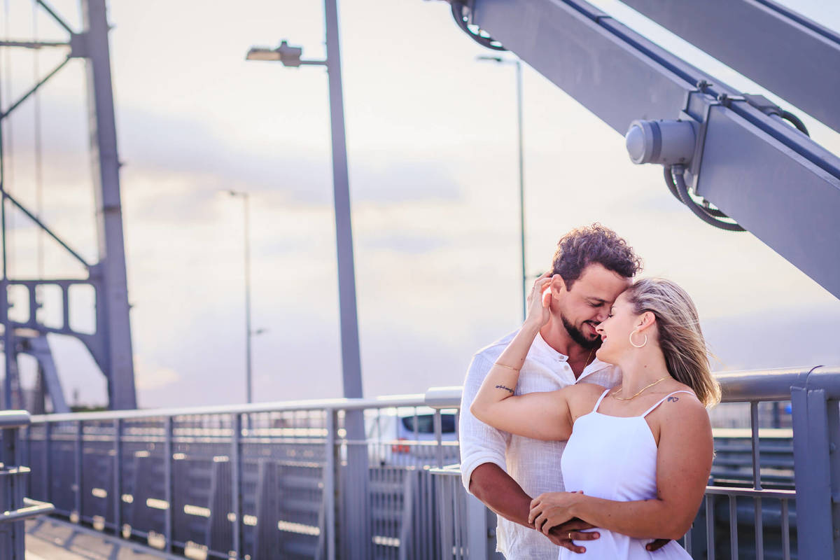 Close do abraço de Bruna e o seu noivo com a Ponte Hercílio Luz iluminada pelo pôr do sol.