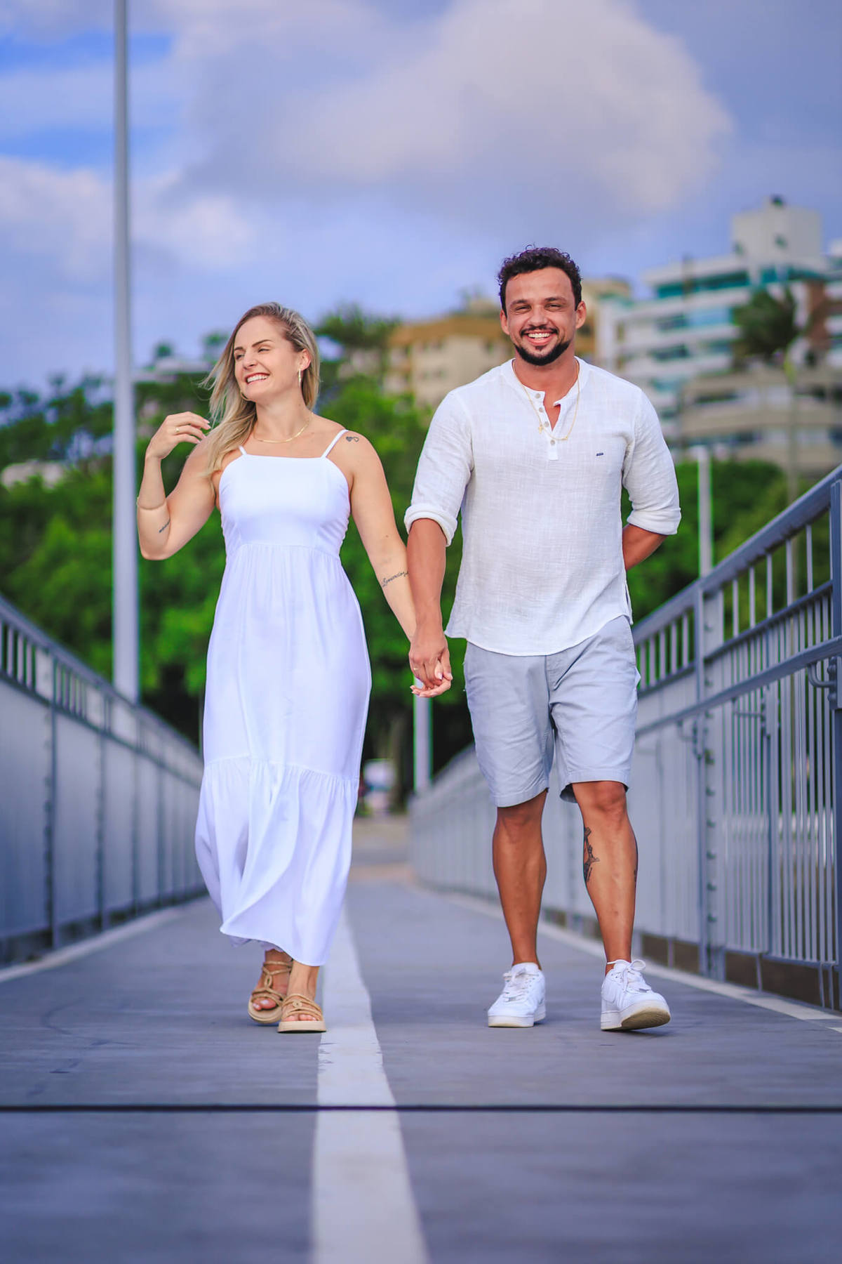 O casal caminha de mãos dadas na cabeceira da Ponte Hercílio Luz, com o mar ao fundo.