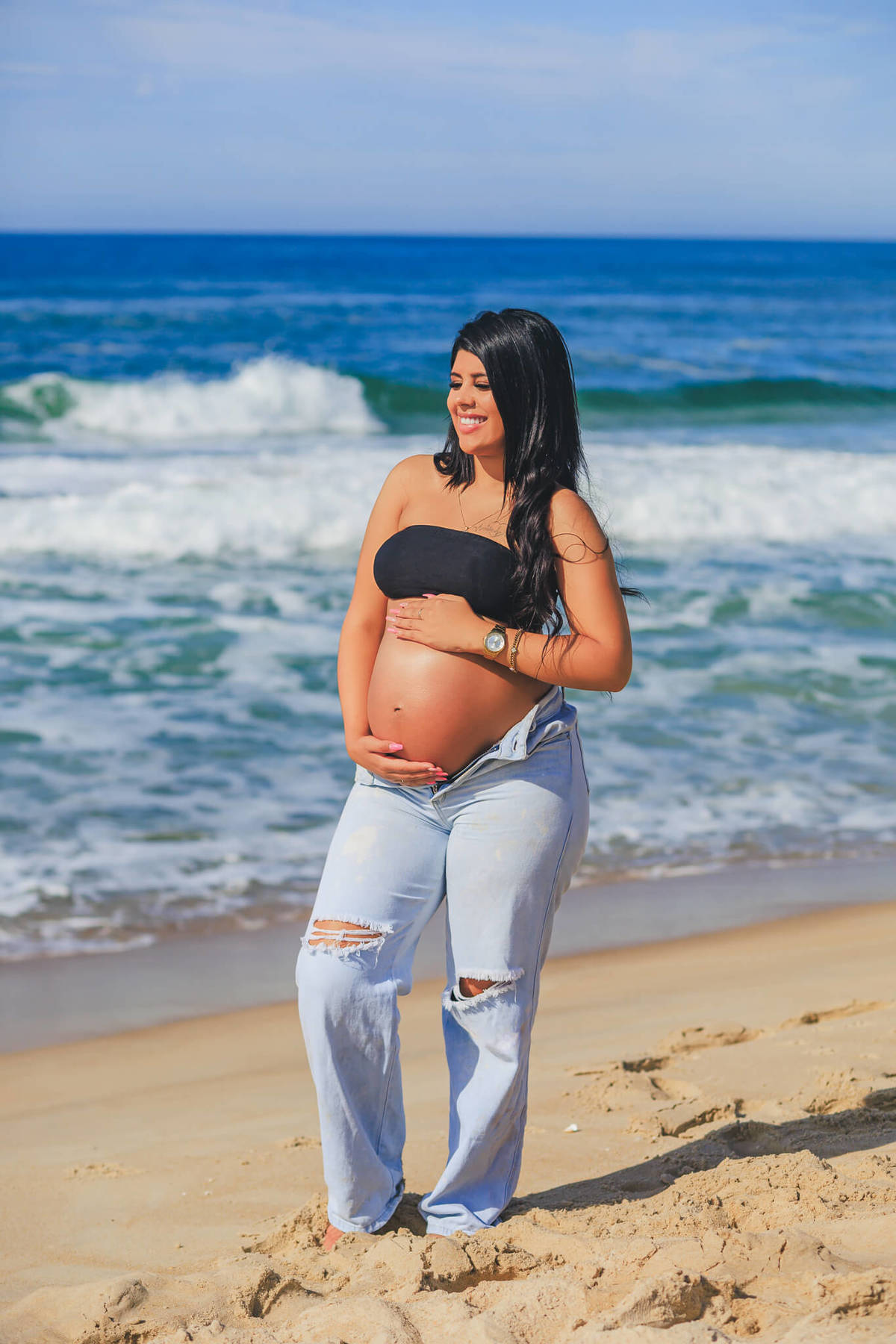 Ensaio gestante na praia em Florianópolis com look casual e luz natural – Maria Eduarda sorrindo e segurando a barriga.