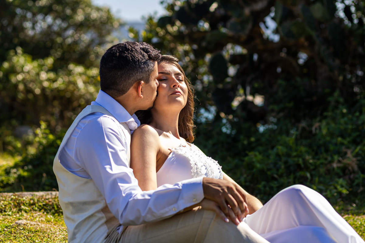 Noiva abraçando o noivo por trás no píer da Praia da Armação, com barcos de pesca ao fundo.