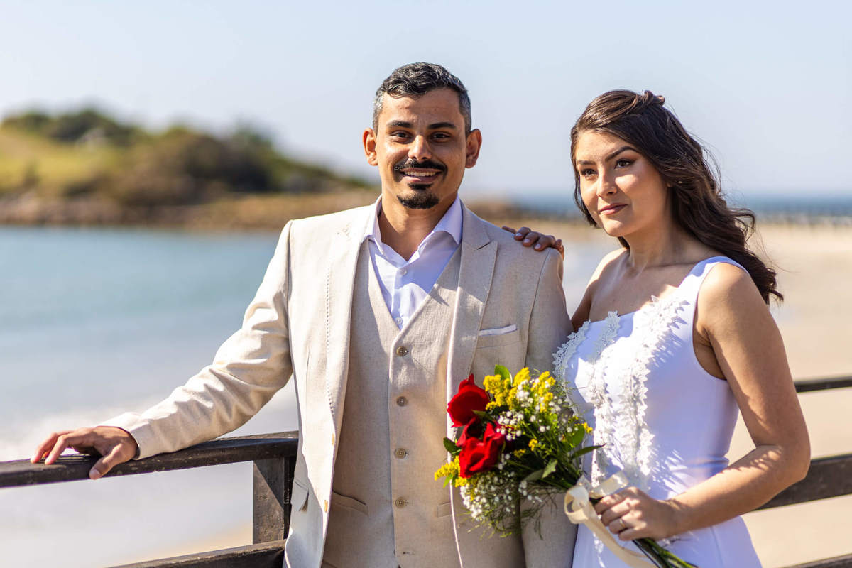 Noivo sorrindo com a noiva ao lado em píer na Praia da Armação durante ensaio de casal em Florianópolis.