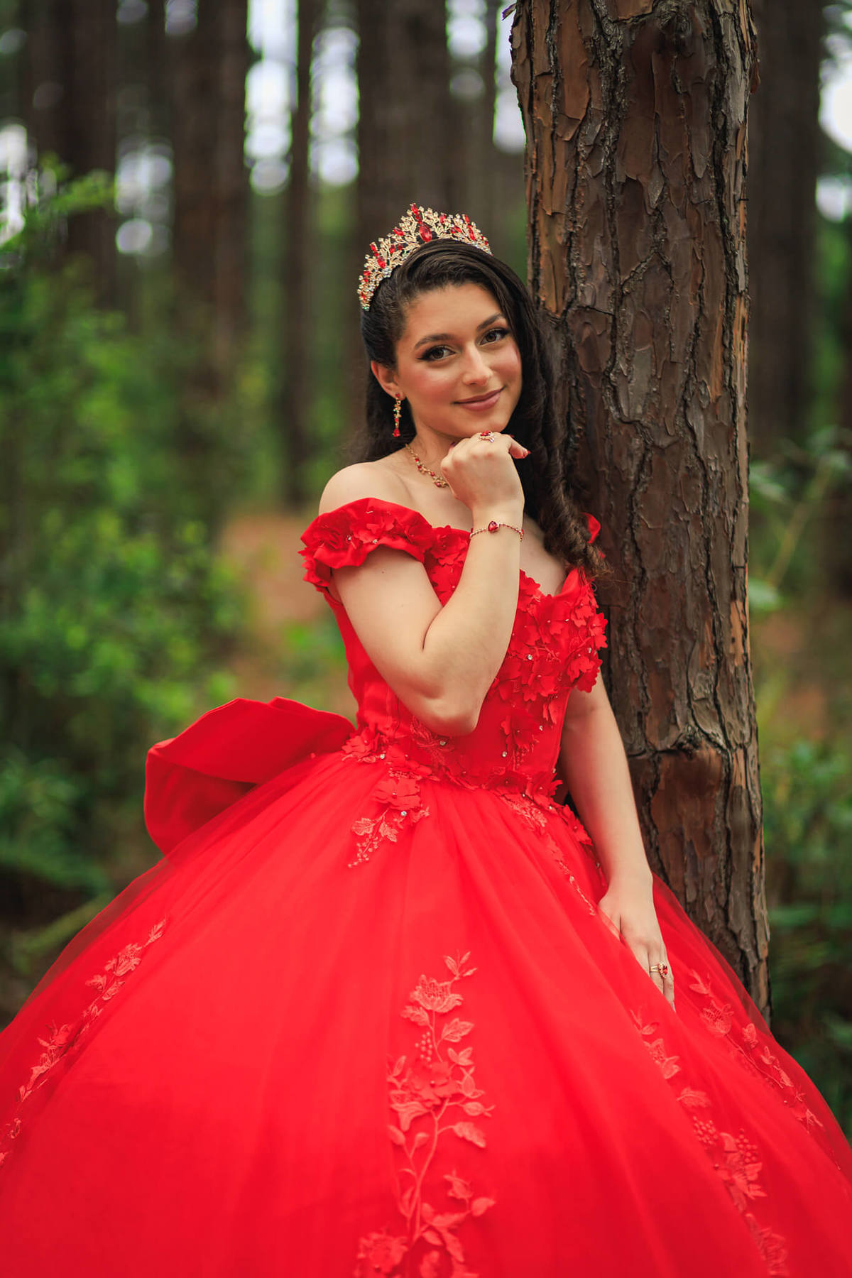 Retrato close-up da debutante encostada em uma árvore no bosque, usando coroa de cristais e vestido vermelho, com fundo desfocado de natureza.
