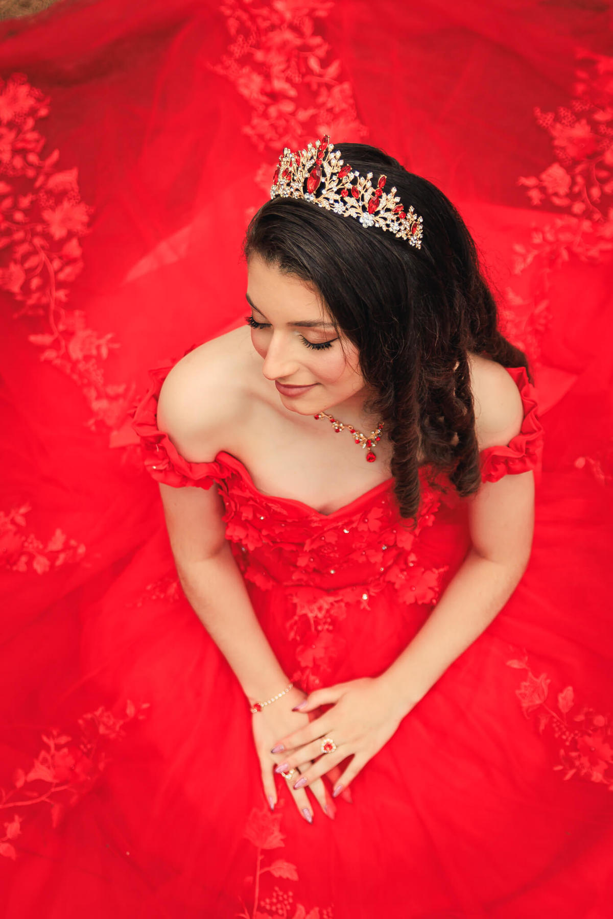 Foto de ângulo superior da debutante sentada no chão da floresta com vestido vermelho rodado espalhado ao redor e olhos fechados.

