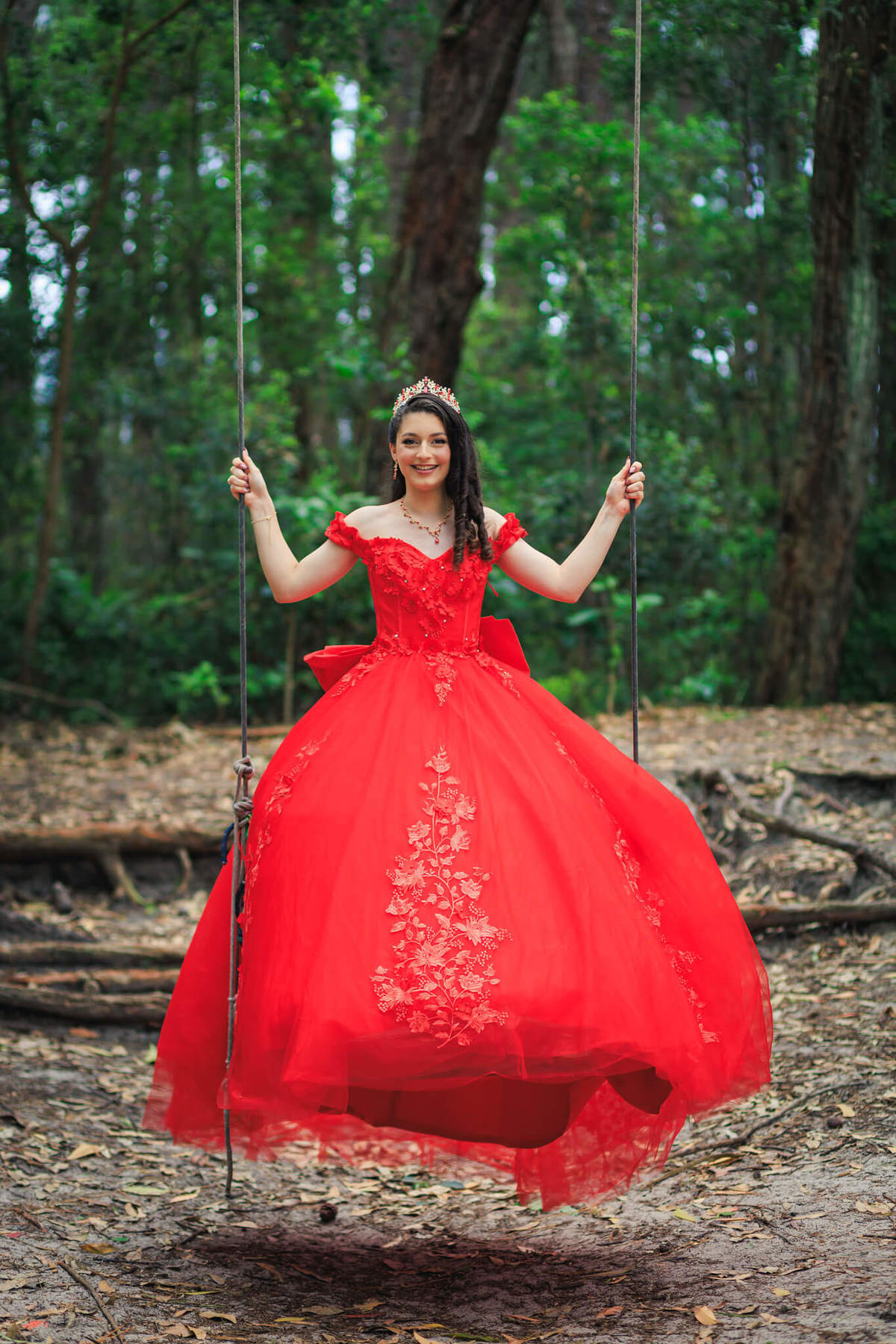 Debutante sorrindo em um balanço rústico na floresta, com vestido de festa vermelho volumoso em movimento.