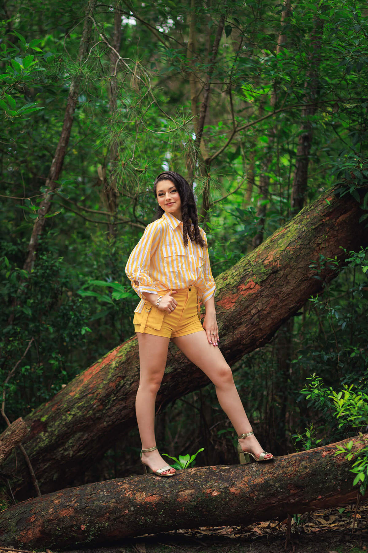 Foto de corpo inteiro da Legna usando short e camisa amarela listrada, em pose espontânea na natureza do Bosque do Moçambique.

