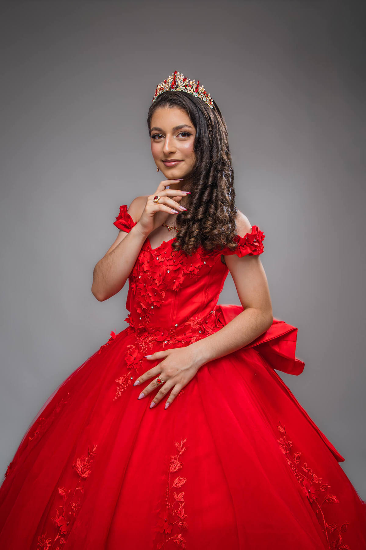 Retrato clássico de 15 anos em estúdio com fundo cinza, debutante usando vestido vermelho bordado e coroa, fazendo pose elegante.
