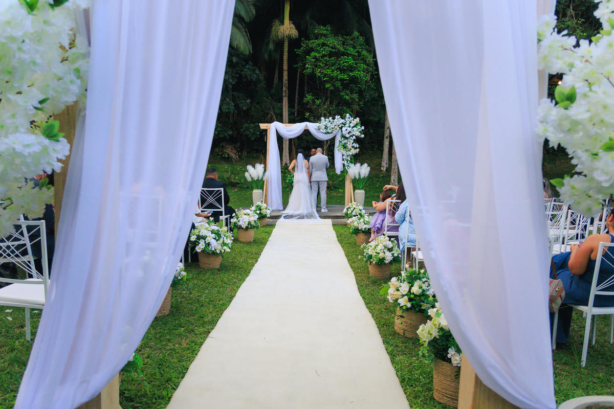 Cerimônia de casamento vista pelo corredor decorado com tecidos brancos e flores.