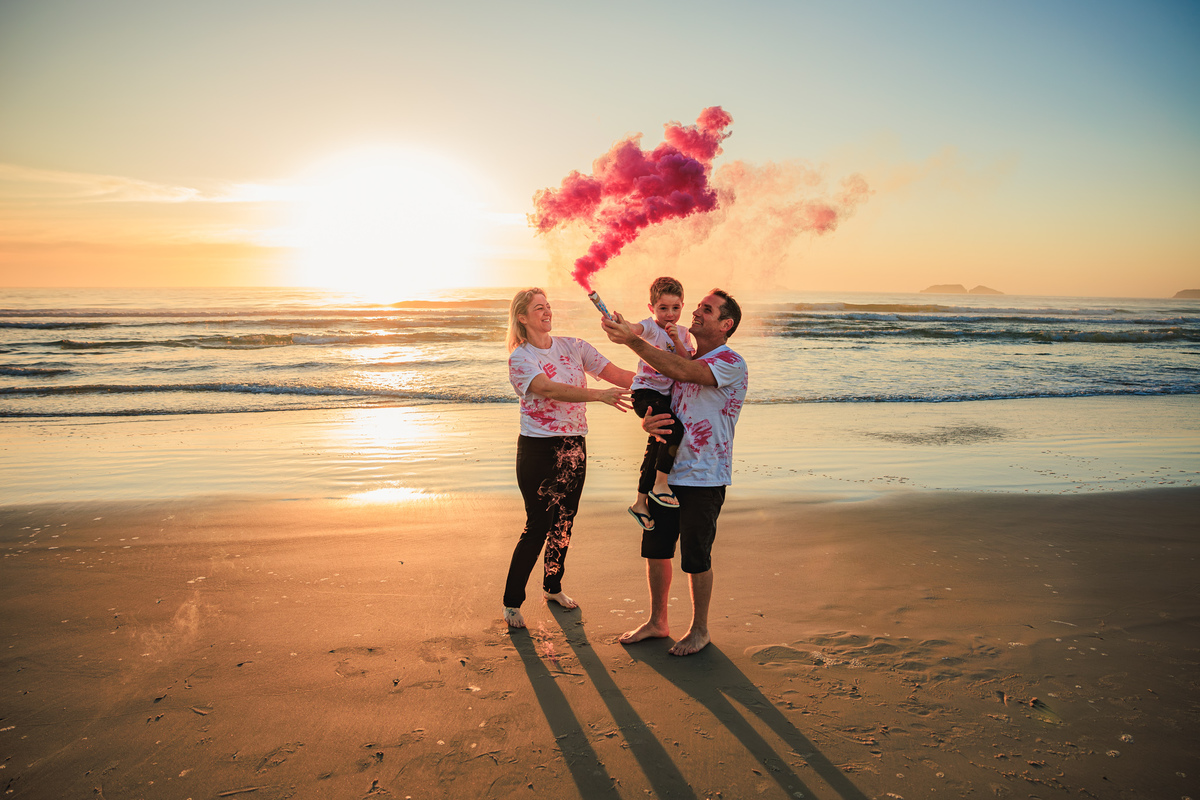 Família celebrando a chegada de uma menina com fumaça rosa na praia.