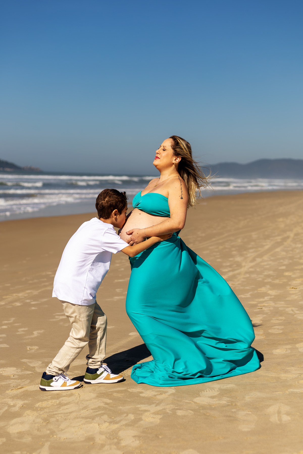 Filho beija a barriga da mãe durante ensaio de gestante na praia em
Florianópolis.