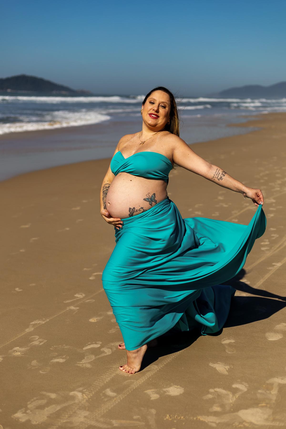 Gestante com vestido azul-turquesa posa à beira-mar durante ensaio
em Florianópolis.