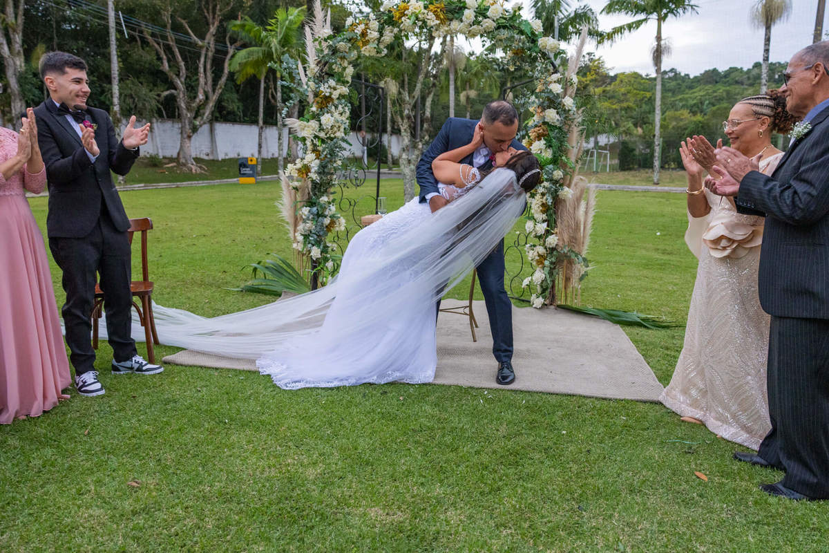 Beijo dos recém-casados diante do arco floral e das pessoas queridas