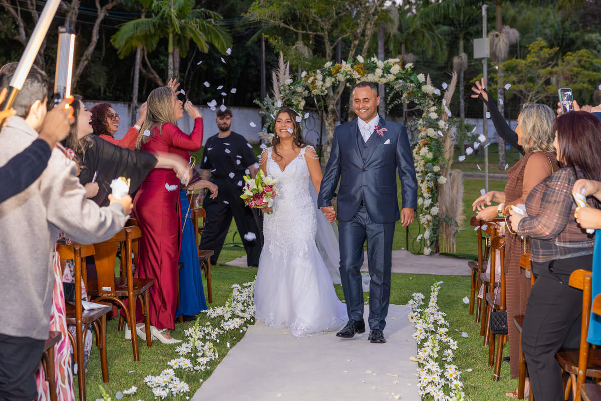 Recém-casados caminham pelo corredor entre pétalas e convidados.