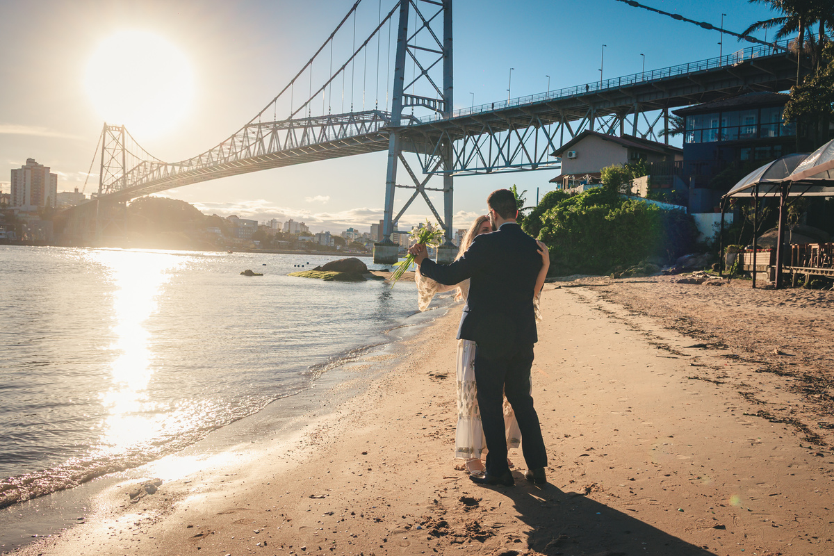 Beijo do casal à beira-mar, com a ponte ao fundo e a luz do fim da tarde.