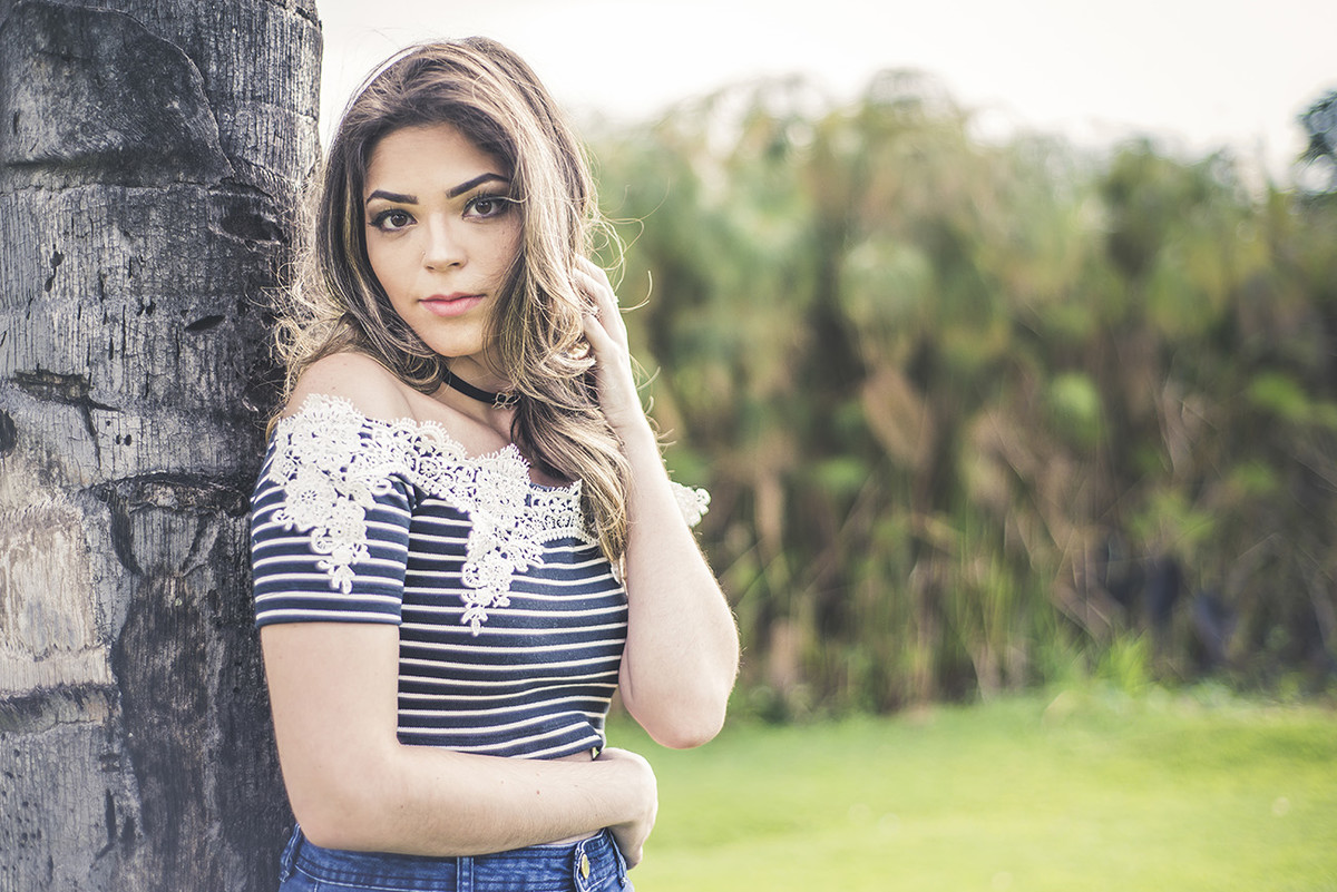 Debutante encostada na árvore passando a mão no cabelo, no jardim do museu de arte da Pampulha em Belo Horizonte BH MG em pose para book de 15 anos