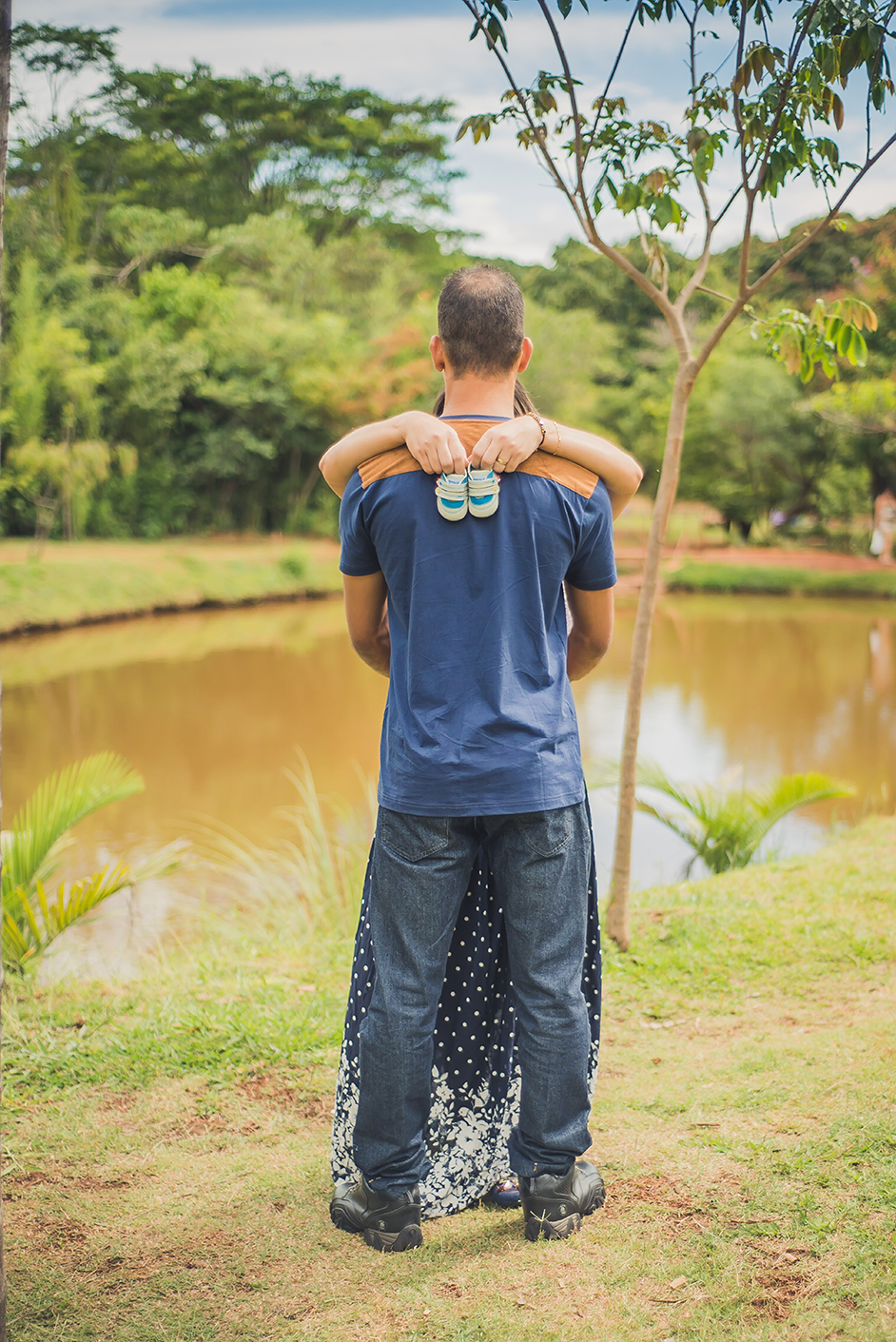 Casal se abraçando próximo a uma lagoa mostrando o sapatinho de bebê enquanto são fotografados pelo fotografo de gestante joão Paulo no parque das águas em belo horizonte BH MG