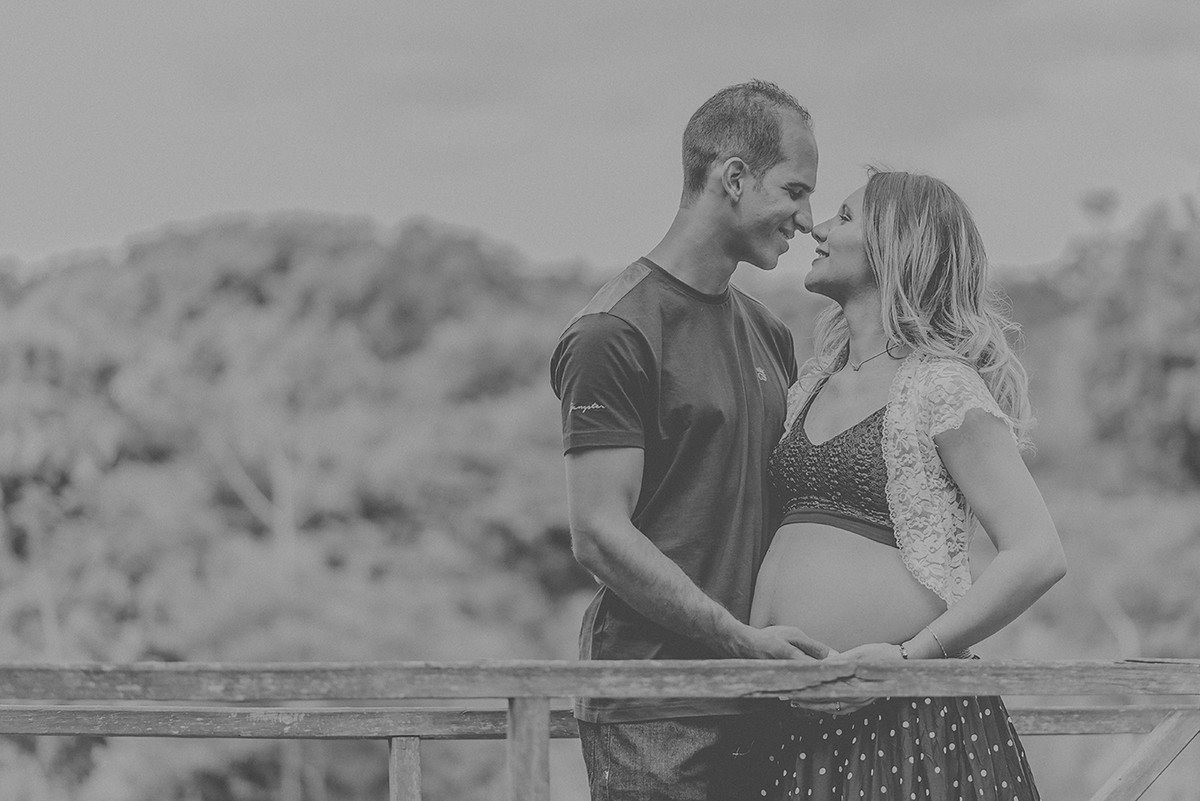 Casal se olhando e com a mão na barriga da gestante em uma ponte de madeira no parque das águas em belo horizonte bh