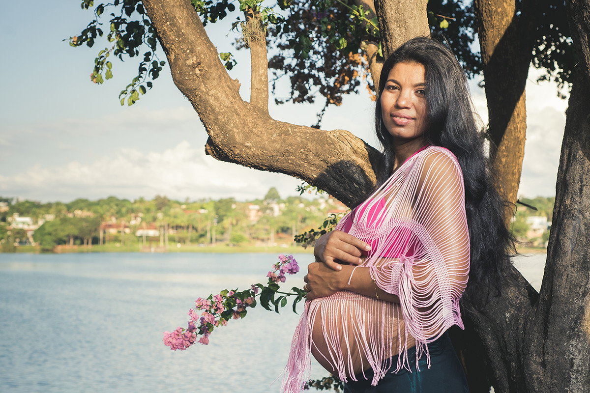 Gestante na lagoa da Pampulha sorrindo próximo a uma árvore com a bela paisagem de belo horizonte BH MG