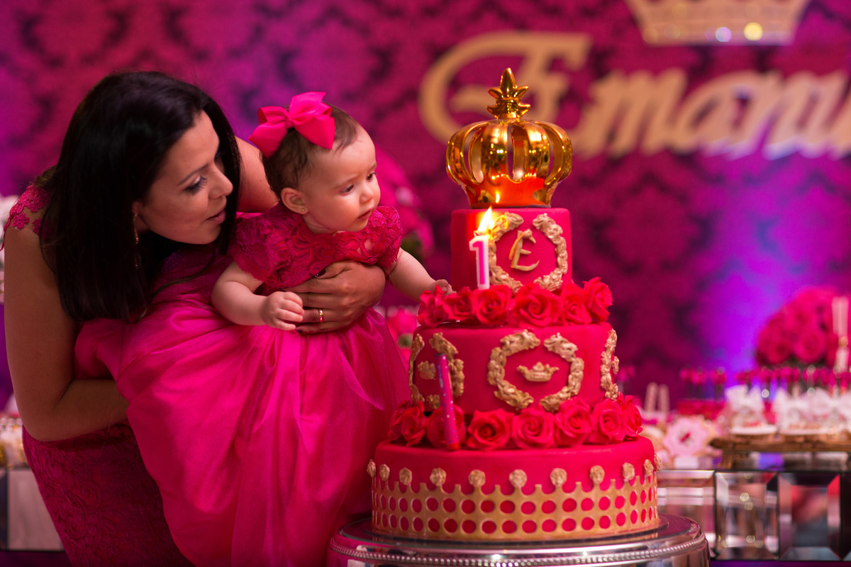 Princesa no colo da mamãe soprando a vela, o bolo vermelho com rosas e a coroa e a vela dão um toque especial