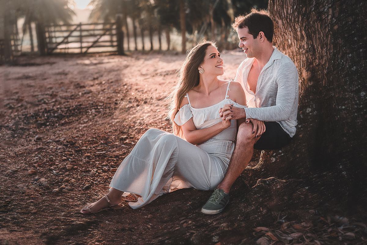 Ensaio_précasamento_Renata_e_Otávio_Ceará-Mirim_Rio_Grande_do_Norte_14_10_2019 Casal sentado na raiz da árvore de mãos dadas, entreolhando-se e sorrindo, enquanto a luz do sol ilumina a imagem em contraluz. 