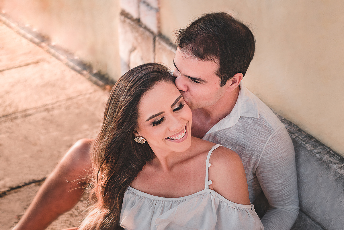 Ensaio_précasamento_Renata_e_Otávio_Ceará-Mirim_Rio_Grande_do_Norte_14_10_2019 Casal sentado no chão. Moça deitada de costas no peito do rapaz, enquanto ele beija seu rosto de forma muito carinhosa, enquanto ela sorri.