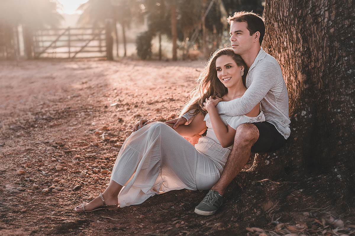 Ensaio_précasamento_Renata_e_Otávio_Ceará-Mirim_Rio_Grande_do_Norte_14_10_2019 Casal sentado na raiz de uma árvore, com as costas encostadas no tronco da árvore. A moça sorri olhando para o horizonte, enquanto o rapaz olha concentrado e a luz do sol entra