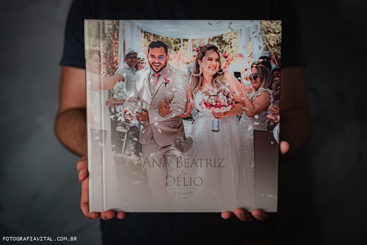 casamento_microwedding_shuibrasil_pureza_riograndedonorte_campo_arlivre_álbum_de_casamento_ Foto da capa do álbum. o Album está sendo segurado por uma pessoa. Na capa vemos uma foto do casal na saída do casamento. Eles sorriem entre as pétalas de rosas.