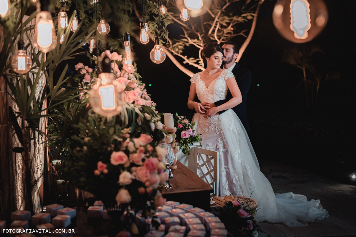 Fazenda Real III - Macaíba - RN - Fotos dos noivos na mesa do bolo. Ela está de costas para ele e ela beija seu rosto entre gambiarras e flores.