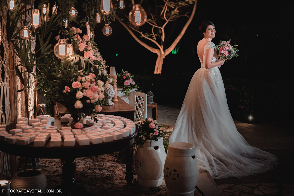 Fazenda Real III - Macaíba - RN - Foto de corpo inteiro da noiva. ela está no meio da decoração. Podemos ver a mesa do bolo, gambiarras e flores. Elas está de costas segurando o buquê e olhando por cima do ombro. 