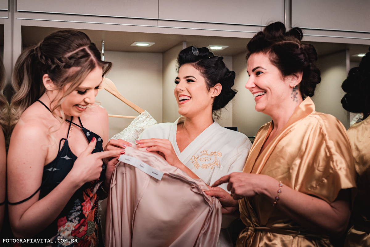 Estúdio Pontes - Natal - RN - Noiva, mãe e amiga da noiva olham a barra do vestido da noiva cheia de nomes e sorriem. 