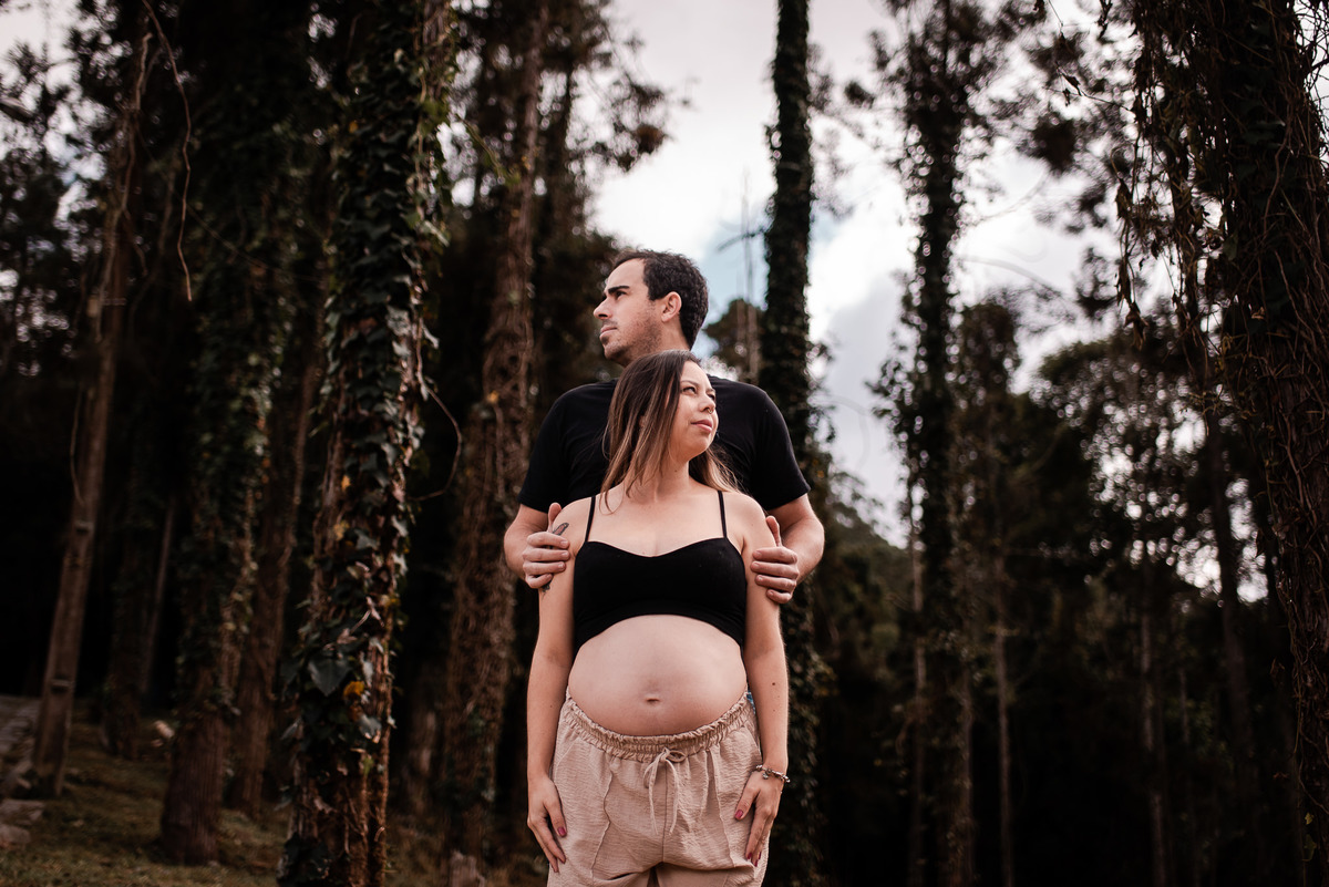 ensaio externo de gestante em nova friburgo, região serrana do rio de janeiro. Foto da gestante de frente olhando para a direita e seu companheiro logo atrás dela com as mãos em seus ombros olhando para o lado oposto