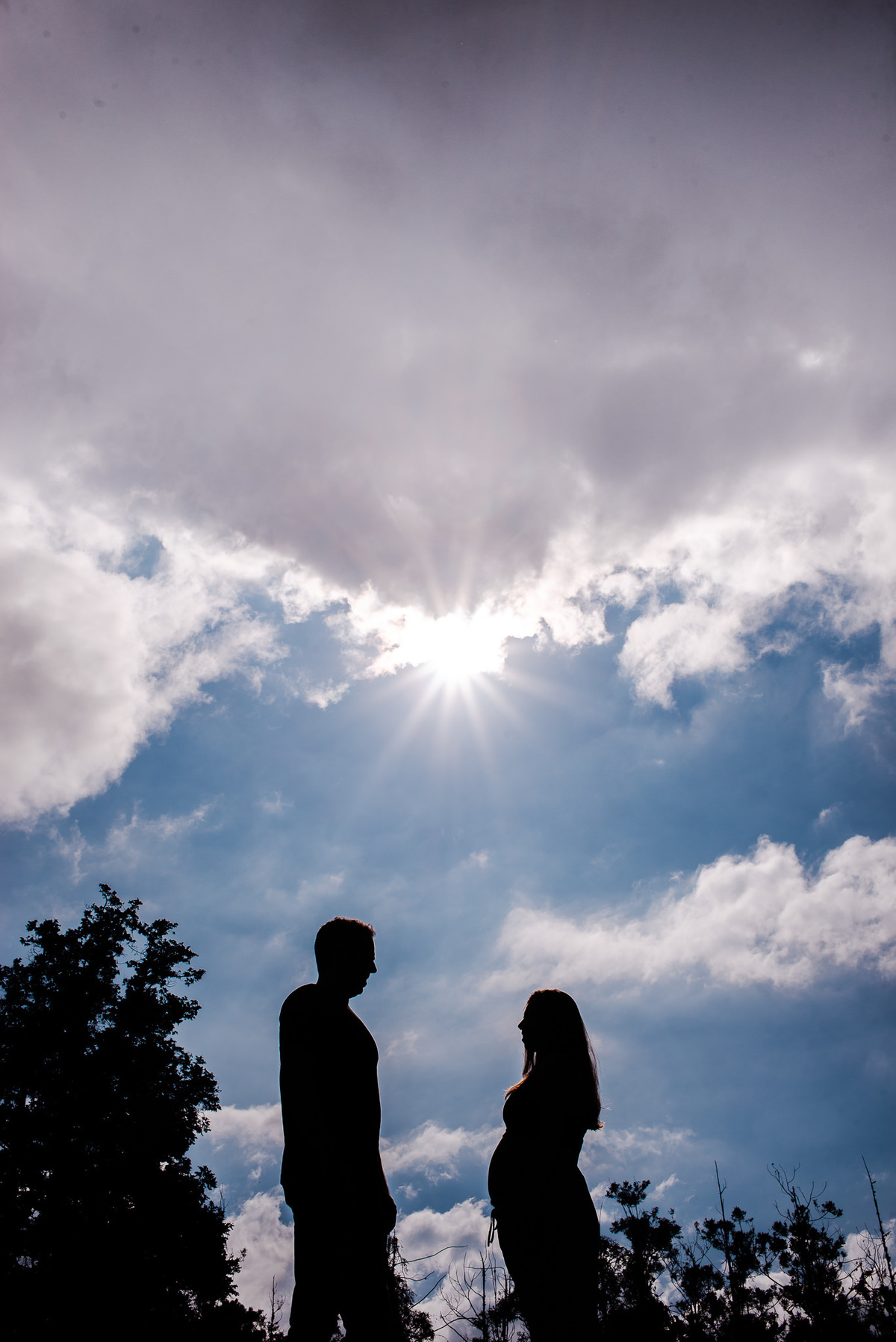 ensaio externo de gestante em nova friburgo, região serrana do rio de janeiro. Foto da silhueta gestante e do companheiro em um parque, mostrando o sol e o céu azul