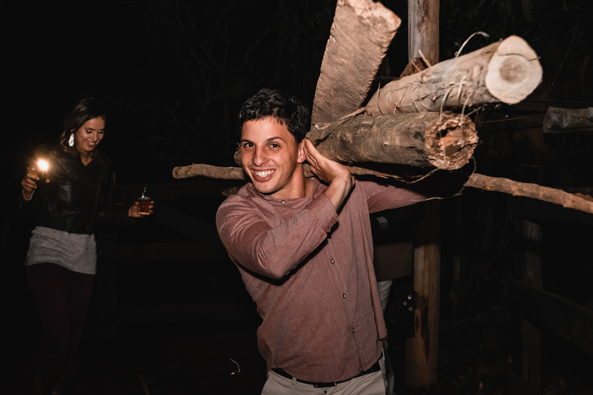 Fotografo de Casamento em Valença - RJ. Casamento em Rio Preto Minas Gerais. Foto da festa de casamento com o convidado carregando madeiras no ombro e a noiva iluminando o caminho com a lanterna do celular