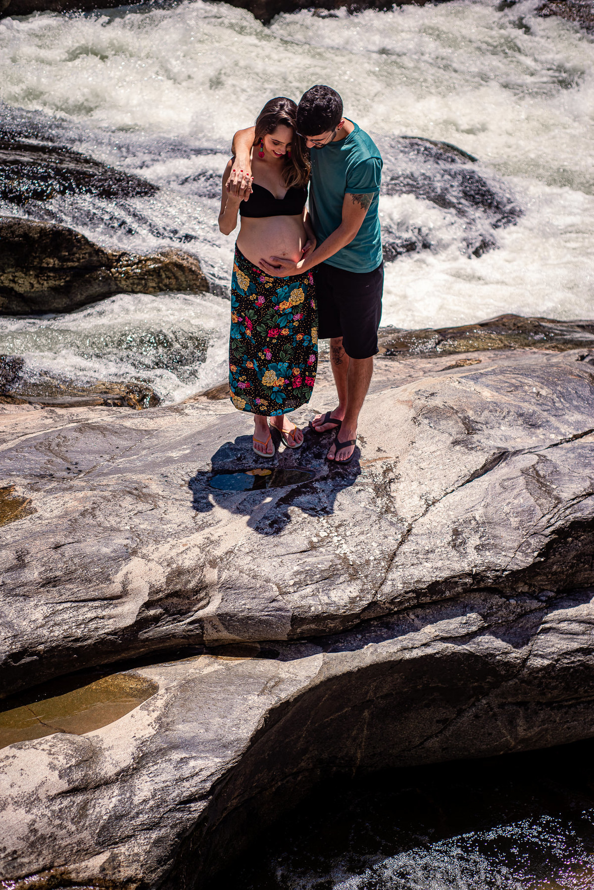 Ensaio externo de gestante na cachoeira em Nova Friburgo, foto da gestante e seu companheiro na pedra de uma cachoeira olhando para a barriga da gestante