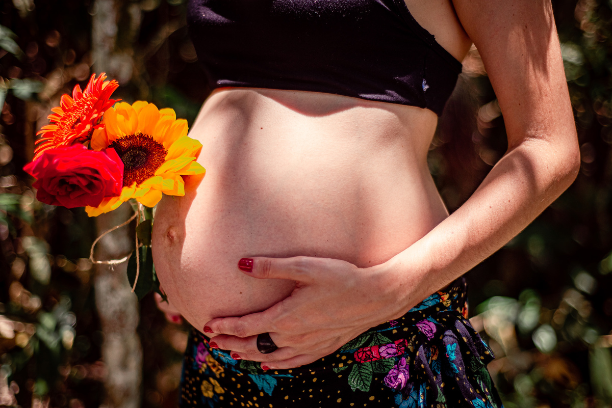 Ensaio externo de gestante na cachoeira em Nova Friburgo, foto da barriga da gestante. A gestante estão com uma das mãos na barriga e a outra segurando flores