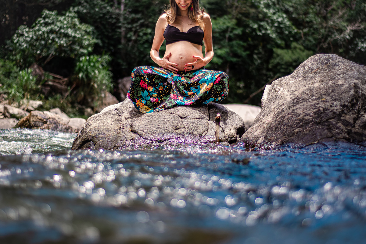 Ensaio externo de gestante na cachoeira em Nova Friburgo, foto da gestante sentada na pedra na cachoeira com as pernas cruzadas olhando para a barriga e com as mãos na barriga
