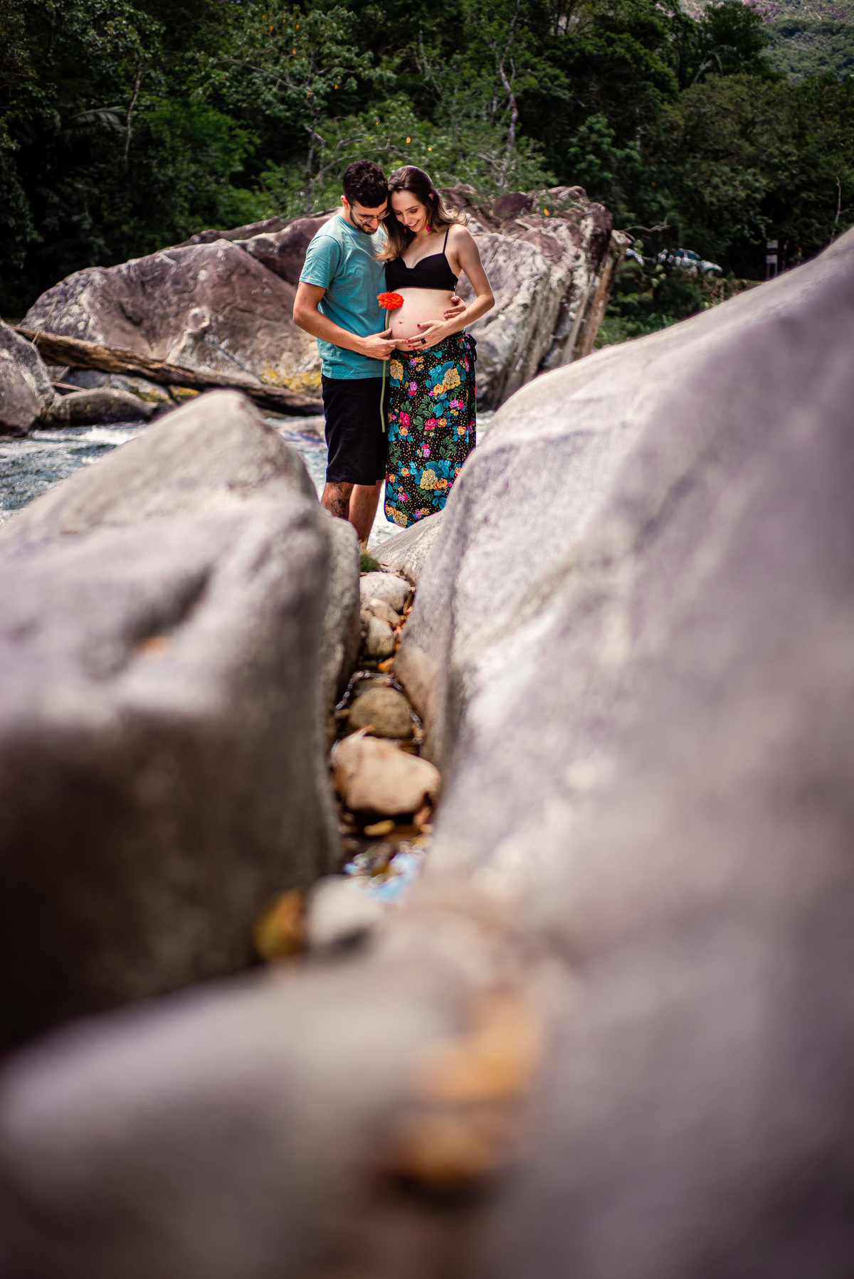 Ensaio externo de gestante em Nova Friburgo, foto feita entre as pedras da cachoeira da gestante e o companheiro segurando uma flor e olhando para a barriga