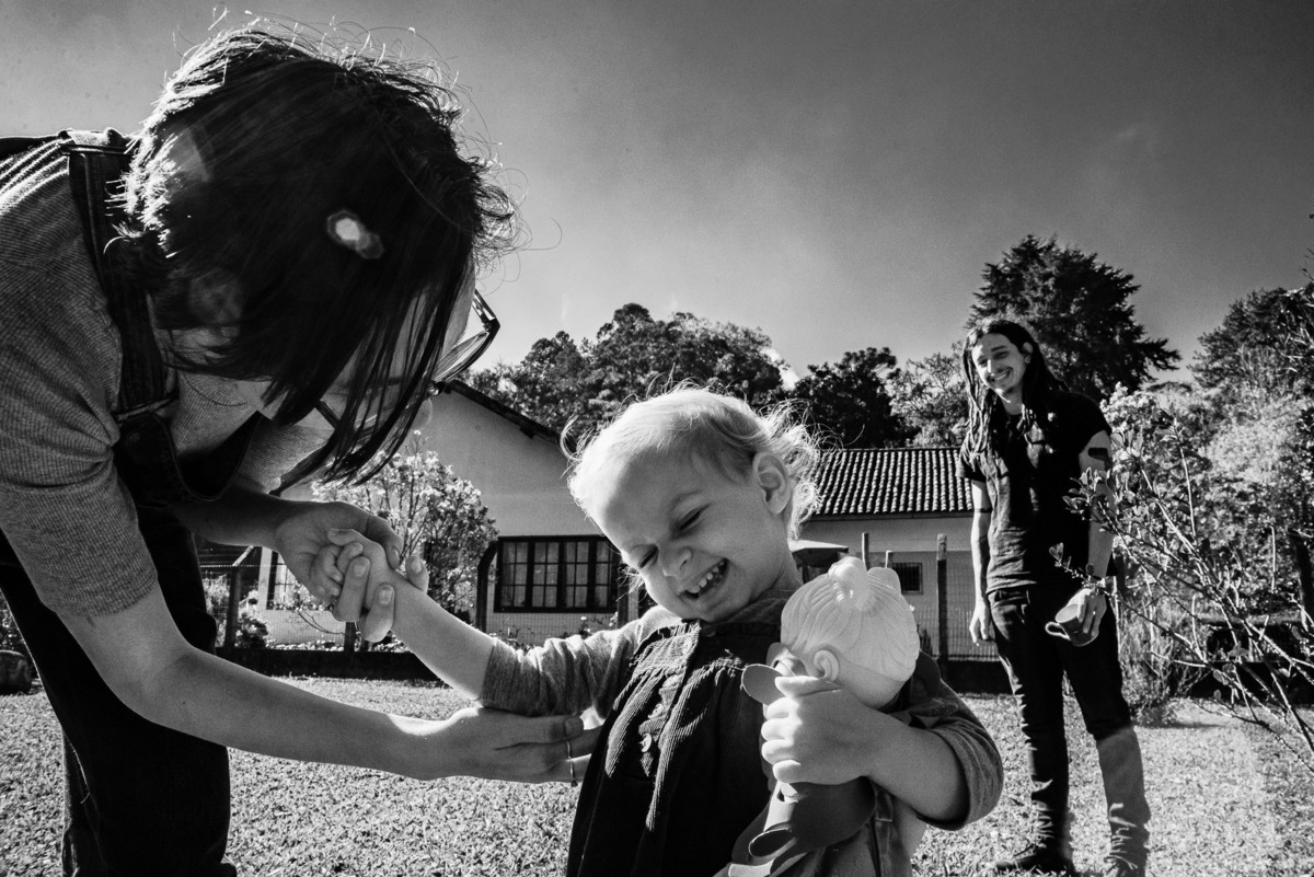 Fotógrafo de família em Nova Friburgo. Ensaio documental de família. Foto em preto e branco da mãe fazendo cócegas na filha e o pai aparecendo ao fundo