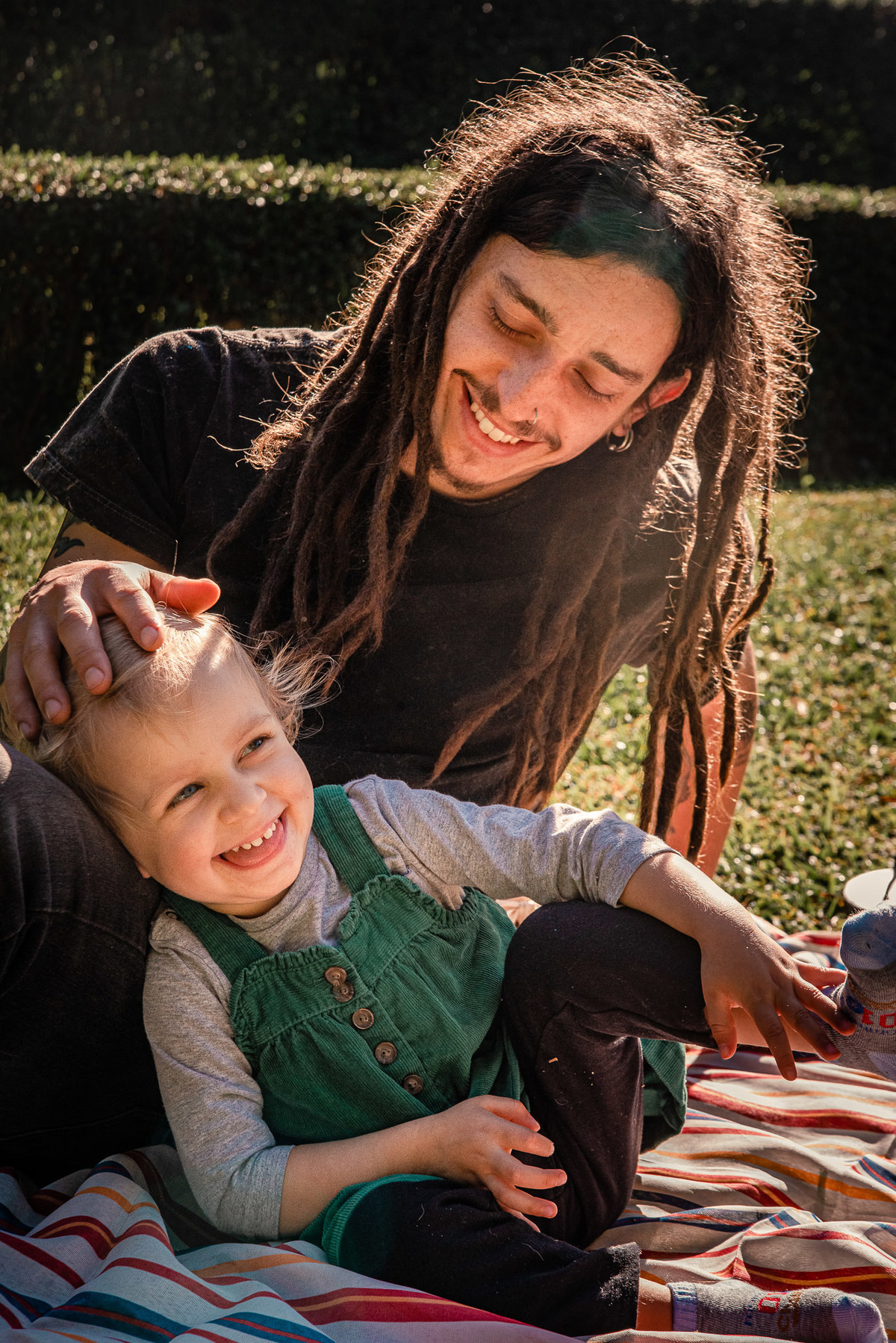 Fotógrafo de família em Nova Friburgo. Ensaio documental de família. Foto da filha sentada com a cabeça apoiada no colo do pai. O pai está fazendo um carinho na criança e os dois estão sorrindo