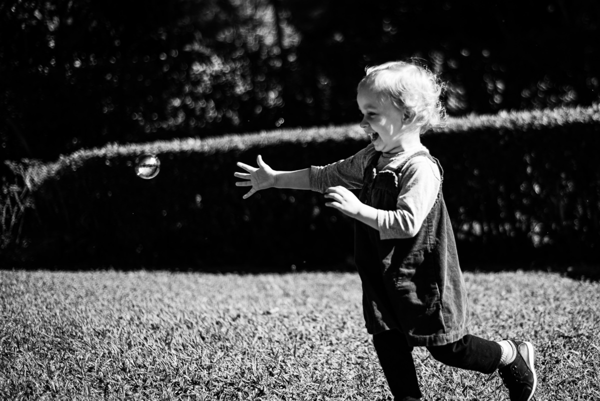 Fotógrafo de família em Nova Friburgo. Ensaio documental de família. Foto em preto e branco da criança correndo no jardim atrás da bolha de sabão