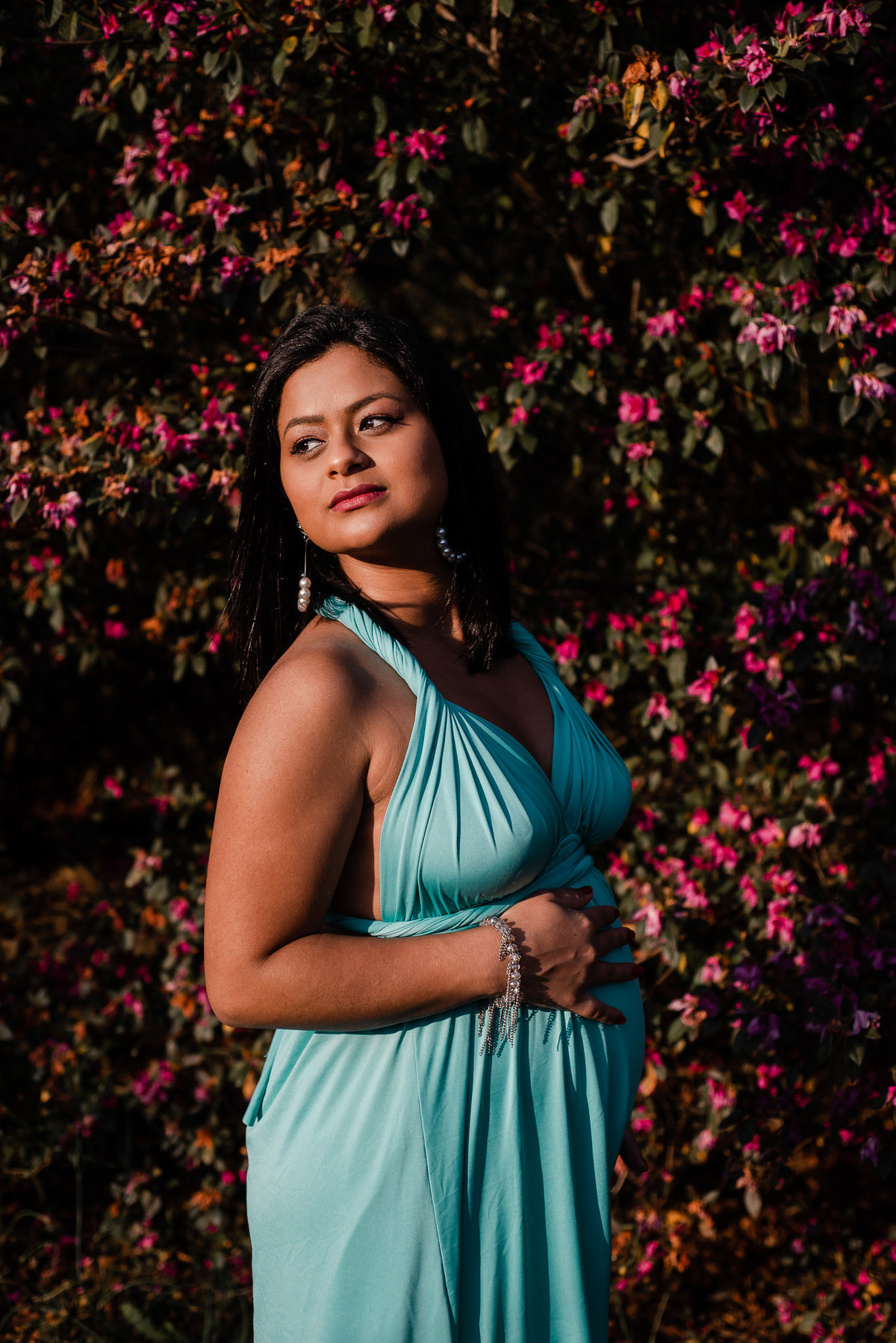 Ensaio externo de gestante em Nova Friburgo, foto da gestante na frente de uma parede de flores com a mão na barriga e olhando por cima do ombro