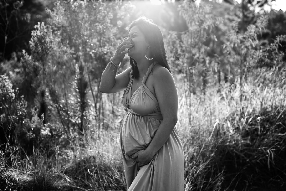 Ensaio externo de gestante em Nova Friburgo, foto em preto e branco da gestante com a mão na barriga rindo na grama alta