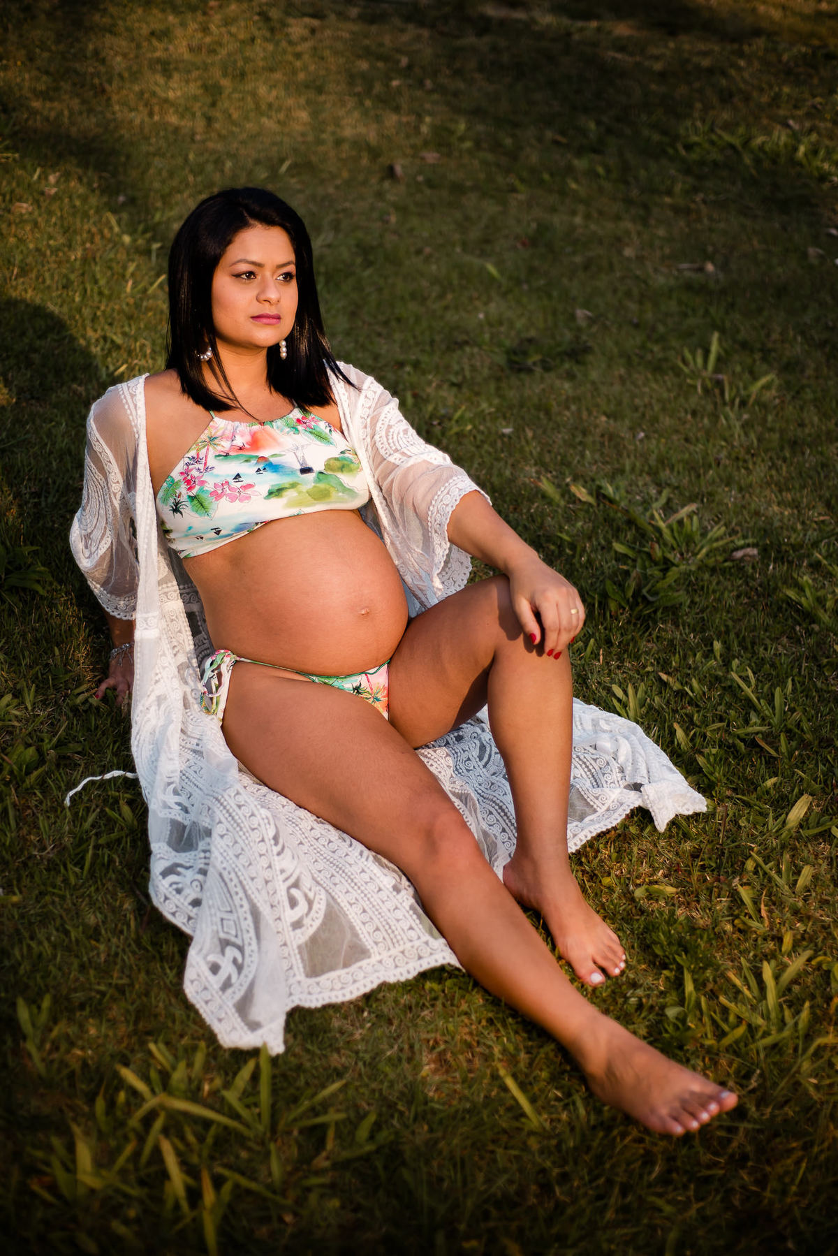 Ensaio externo de gestante em Nova Friburgo, foto da gestante sentada na grama com uma das mãos apoiada no chão e a outra sob o joelho 