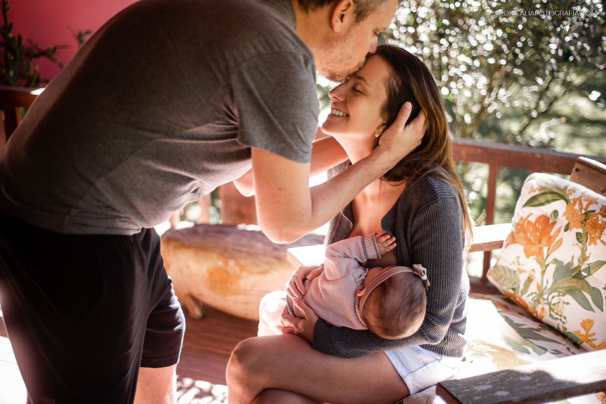 Ensaio documental de família em casa feito por fotógrafos em nova friburgo região serrana do rio de janeiro. Foto da mãe sentada com a filha no colo e o pai dando um beijo na teta da esposa.