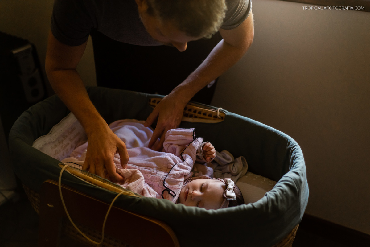 Ensaio documental de família em casa feito por fotógrafos em nova friburgo região serrana do rio de janeiro. Foto do pai cobrindo a filha no berço