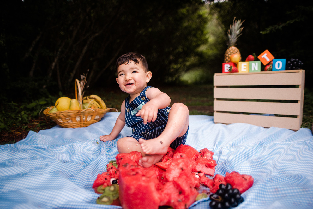 Fotógrafo de família em Nova Friburgo. Ensaio smash the cake da criança. Foto da criança sentada rindo atrás do bolo de melancia destruído