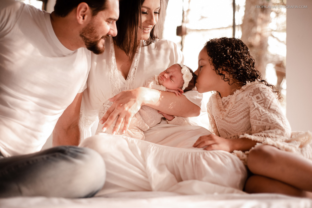 Ensaio documental de família em casa feito por fotógrafos em nova friburgo região serrana do rio de janeiro. Foto da família sentada na cama, com a filha mais velha dando um beijo na cabeça da irmã recém nascida que está no colo da mãe
