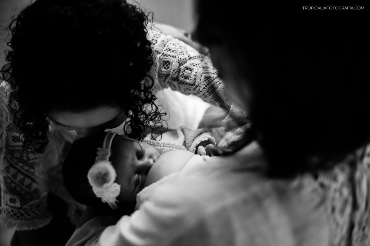 Ensaio documental de família em casa feito por fotógrafos em nova friburgo região serrana do rio de janeiro. Foto em preto e branco da mãe amamentando e a filha mais velha dando um beijo na cabeça do bebê